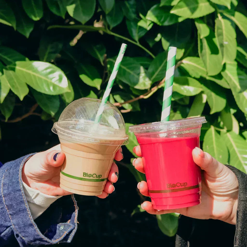 Two hands holding takeaway cups against a leafy green background. One contains an iced latte with a dome lid, and the other a vibrant red smoothie. Both feature the green BioCup logo and striped paper straws.