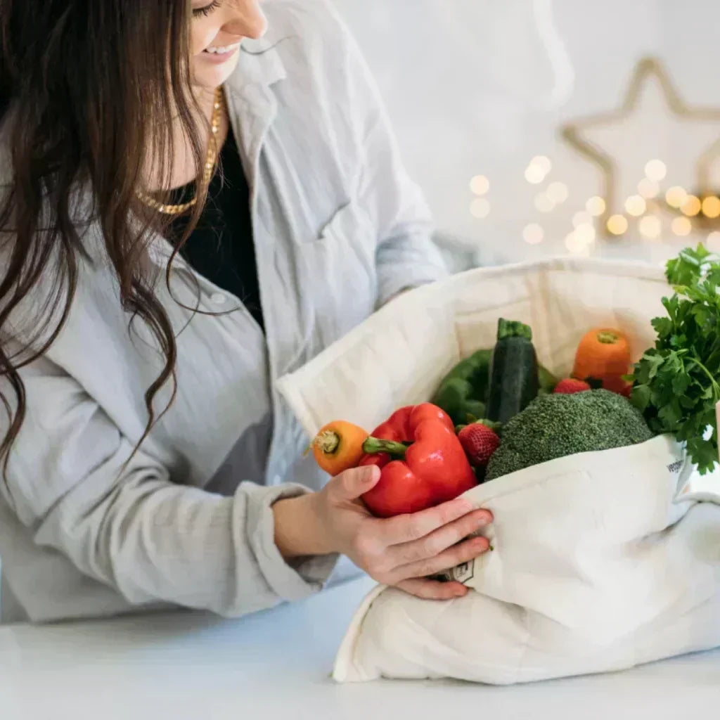 Smiling woman holding a reusable canvas bag filled with fresh produce like red capsicum, broccoli, and carrots.
