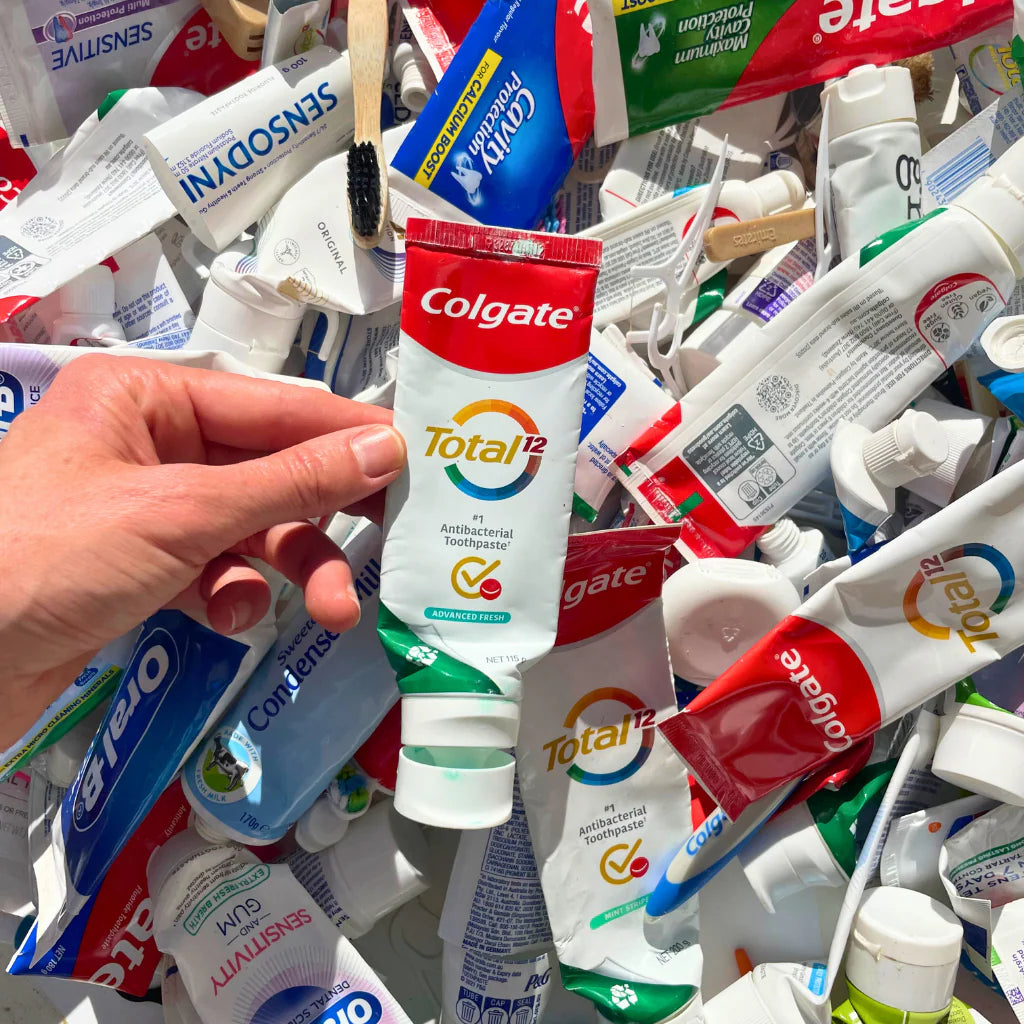 Hand holding a Colgate toothpaste tube amidst a pile of various brand toothpaste tubes.